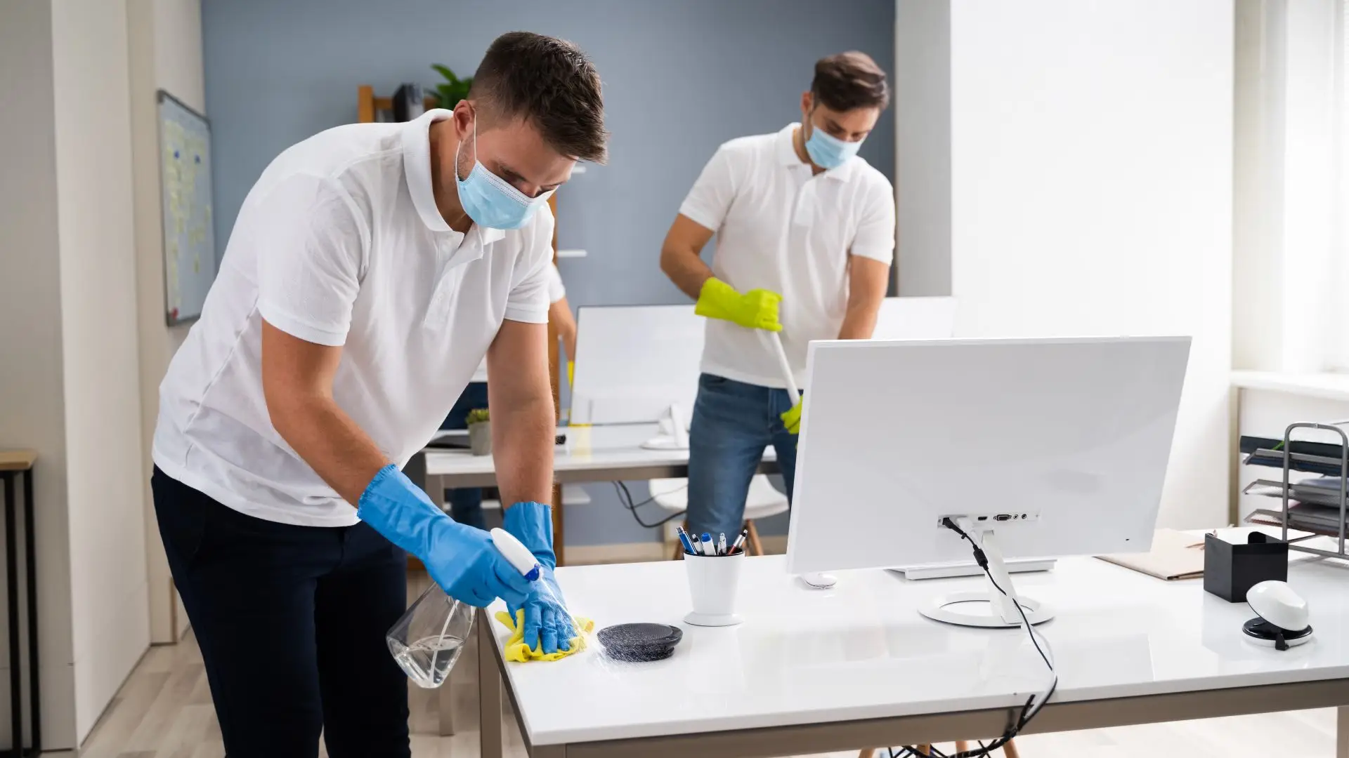 Two men in white shirts and blue gloves cleaning a desk in an office environment.
